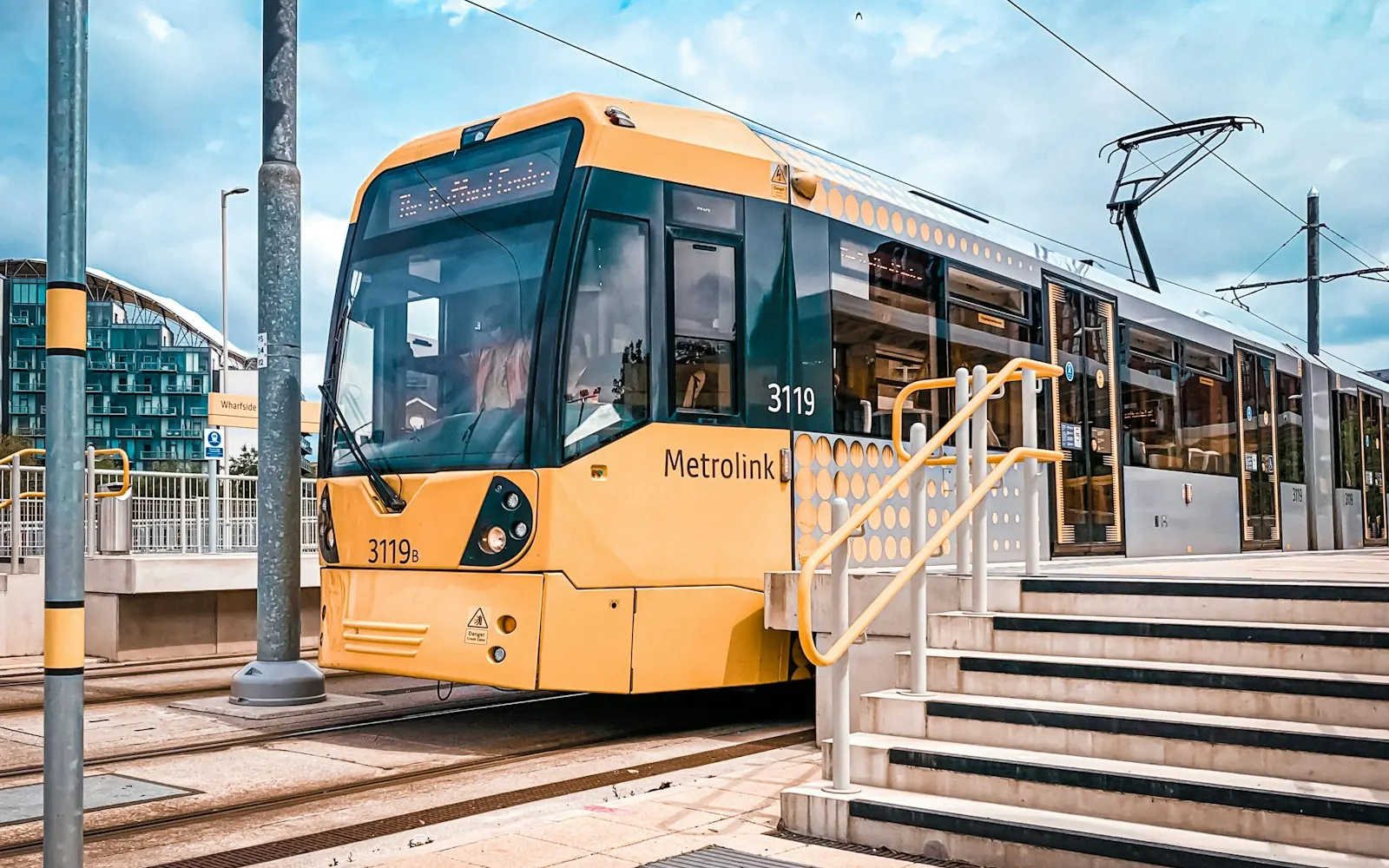 A yellow Metrolink tram at a stop in Manchester, where many people browse on their phones