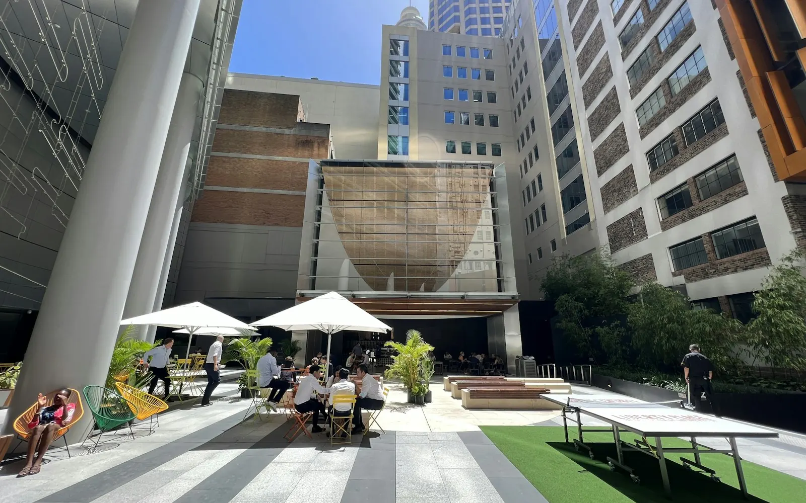 Office workers at outdoor tables under umbrellas in a Sydney CBD plaza surrounded by towers