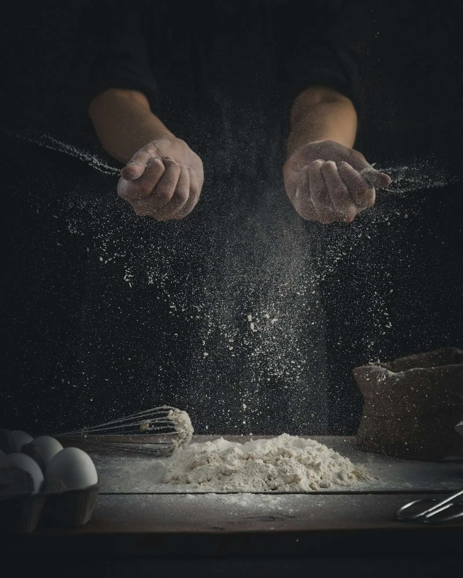 Flour being poured onto a dark table beside eggs and a whisk